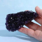 Hand holding a dark purple amethyst crystal formation against a light blue background